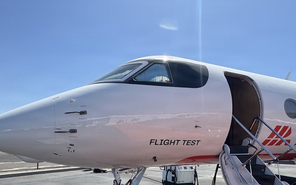 A white business jet marked as a 'Flight Test' aircraft belonging to Honeywell Aerospace is parked on an airport tarmac. The aircraft's door is open with boarding stairs deployed, and ground support equipment is visible beneath the fuselage. The livery features red and white colors with the Honeywell Aerospace logo and branding prominently displayed.
