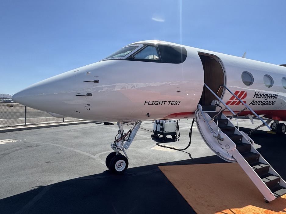 A white business jet marked as a 'Flight Test' aircraft belonging to Honeywell Aerospace is parked on an airport tarmac. The aircraft's door is open with boarding stairs deployed, and ground support equipment is visible beneath the fuselage. The livery features red and white colors with the Honeywell Aerospace logo and branding prominently displayed.