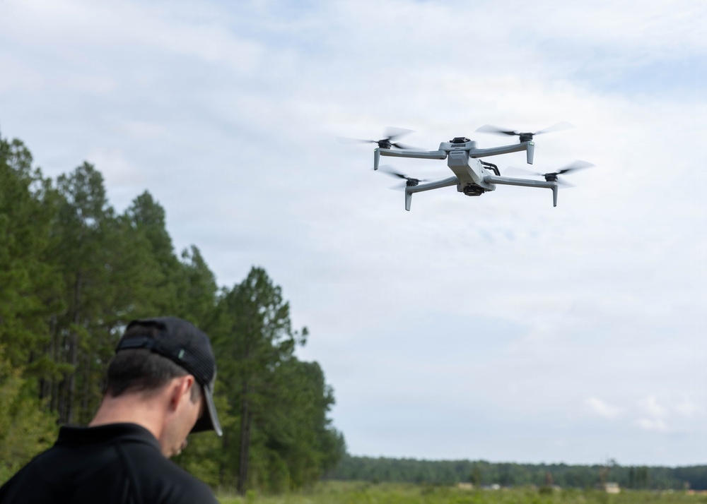 A man wearing a black cap and dark shirt is operating a quadcopter drone in an outdoor setting. The drone, a gray consumer-grade quadcopter with a camera gimbal, is hovering in the air with its propellers visibly spinning. The background features a treeline of tall pine trees under a partly cloudy sky.