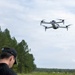 A man wearing a black cap and dark shirt is operating a quadcopter drone in an outdoor setting. The drone, a gray consumer-grade quadcopter with a camera gimbal, is hovering in the air with its propellers visibly spinning. The background features a treeline of tall pine trees under a partly cloudy sky.