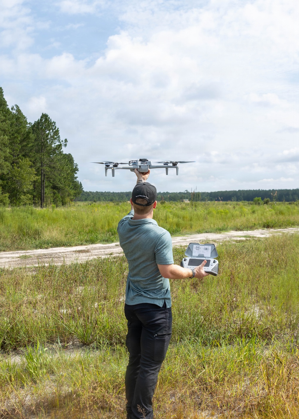 A man is standing in an open grassy field preparing to launch a quadcopter drone by holding it overhead with one hand while operating a remote controller with the other. The setting appears to be a rural or semi-wild area with pine trees and scrubland in the background. The image is well-lit with natural daylight and partially cloudy skies.