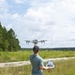 A man is standing in an open grassy field preparing to launch a quadcopter drone by holding it overhead with one hand while operating a remote controller with the other. The setting appears to be a rural or semi-wild area with pine trees and scrubland in the background. The image is well-lit with natural daylight and partially cloudy skies.