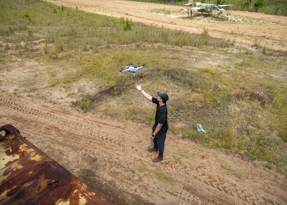 A person dressed in all black is operating or catching a small quadcopter drone in an open sandy field. In the background, a decommissioned military aircraft painted in camouflage colors is visible on the ground. The scene appears to be at a military training area or abandoned airfield.