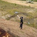 A person dressed in all black is operating or catching a small quadcopter drone in an open sandy field. In the background, a decommissioned military aircraft painted in camouflage colors is visible on the ground. The scene appears to be at a military training area or abandoned airfield.