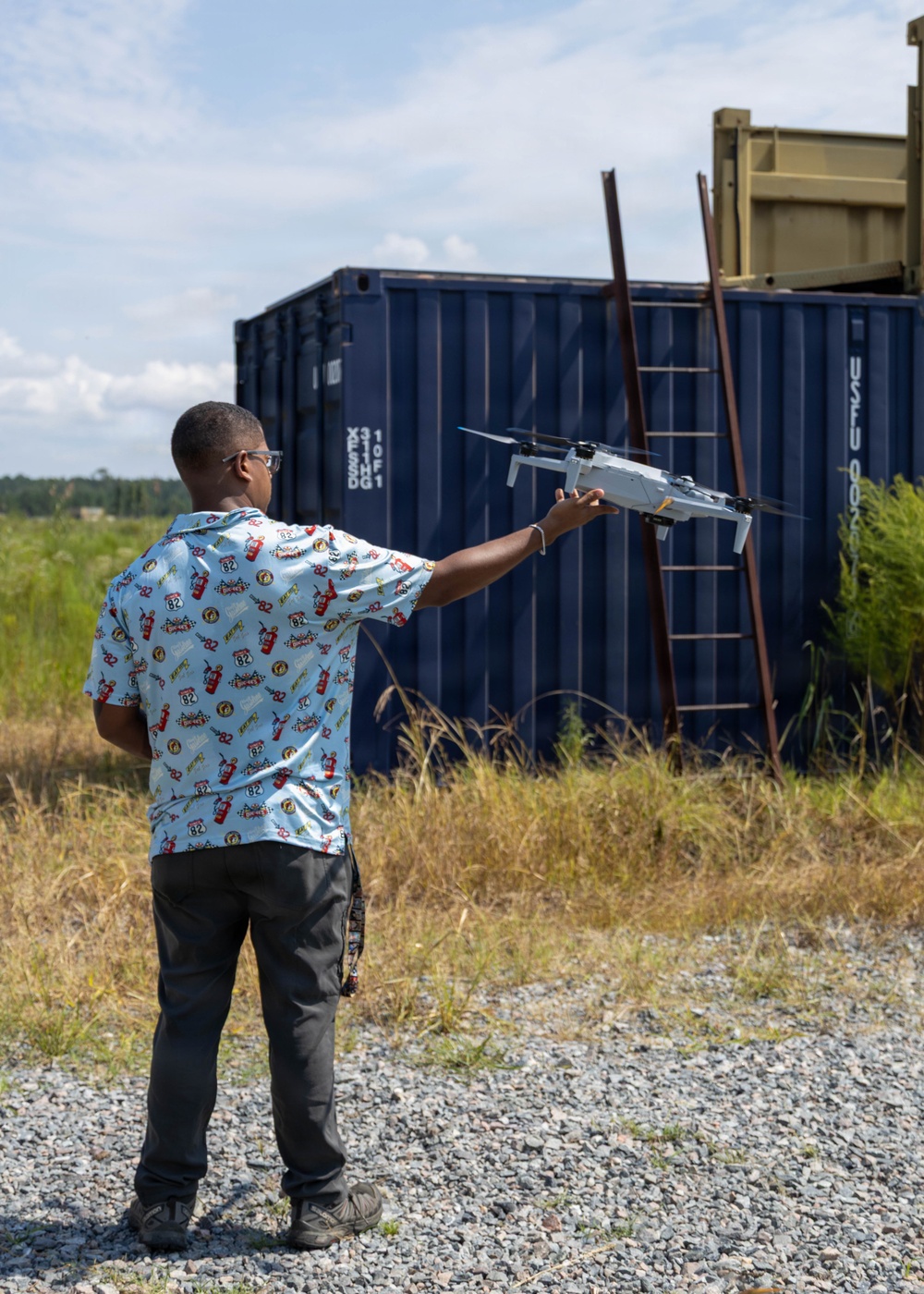 A person is holding a small consumer drone (appears to be a DJI model) outstretched in one hand, likely preparing to launch or having just caught it. The scene takes place outdoors on a gravel surface near blue shipping containers and a metal ladder structure. The setting appears to be an open field or training/industrial area with vegetation in the background.