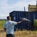 A person is holding a small consumer drone (appears to be a DJI model) outstretched in one hand, likely preparing to launch or having just caught it. The scene takes place outdoors on a gravel surface near blue shipping containers and a metal ladder structure. The setting appears to be an open field or training/industrial area with vegetation in the background.