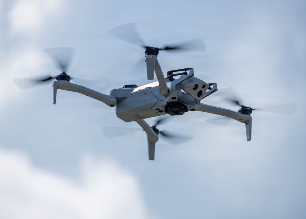 A consumer-grade quadcopter drone is captured mid-flight against an overcast sky. The drone is light gray in color and features four spinning propellers, extended landing legs, and a visible camera gimbal system mounted on its underside. The image is taken from a slightly below-angle perspective, providing a clear view of the drone's undercarriage and sensor array.