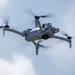 A consumer-grade quadcopter drone is captured mid-flight against an overcast sky. The drone is light gray in color and features four spinning propellers, extended landing legs, and a visible camera gimbal system mounted on its underside. The image is taken from a slightly below-angle perspective, providing a clear view of the drone's undercarriage and sensor array.