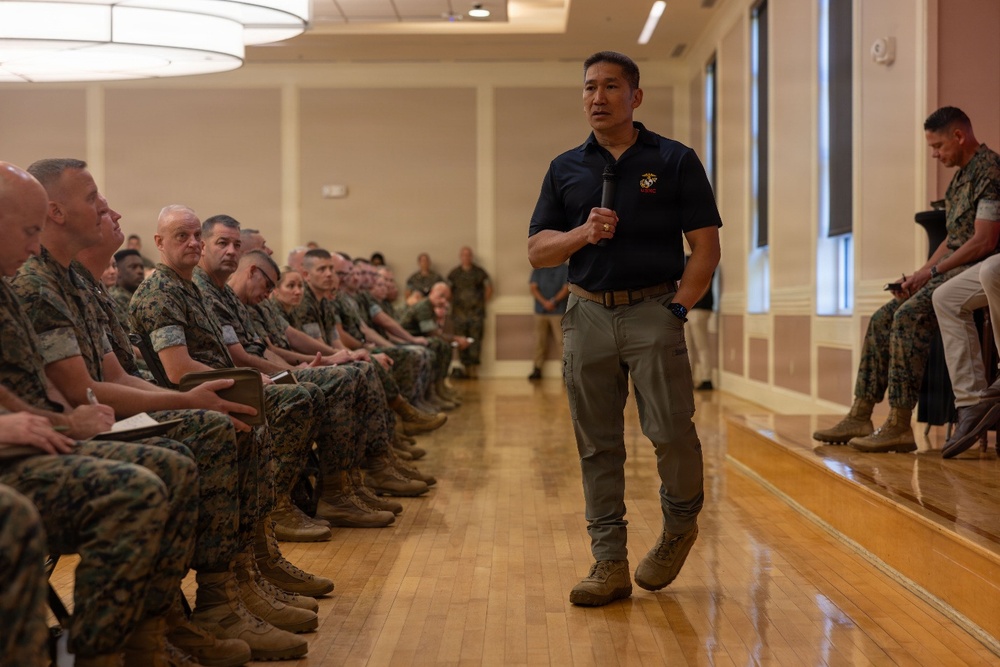 A civilian or senior official wearing a navy blue polo shirt with a Marine Corps emblem is addressing a large group of U.S. Marines in MARPAT camouflage uniforms seated in rows in a formal hall. The speaker is holding a microphone and appears to be walking while presenting, suggesting a briefing, lecture, or town hall-style event. The setting appears to be a military installation meeting or conference room with polished hardwood floors.