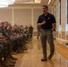 A civilian or senior official wearing a navy blue polo shirt with a Marine Corps emblem is addressing a large group of U.S. Marines in MARPAT camouflage uniforms seated in rows in a formal hall. The speaker is holding a microphone and appears to be walking while presenting, suggesting a briefing, lecture, or town hall-style event. The setting appears to be a military installation meeting or conference room with polished hardwood floors.