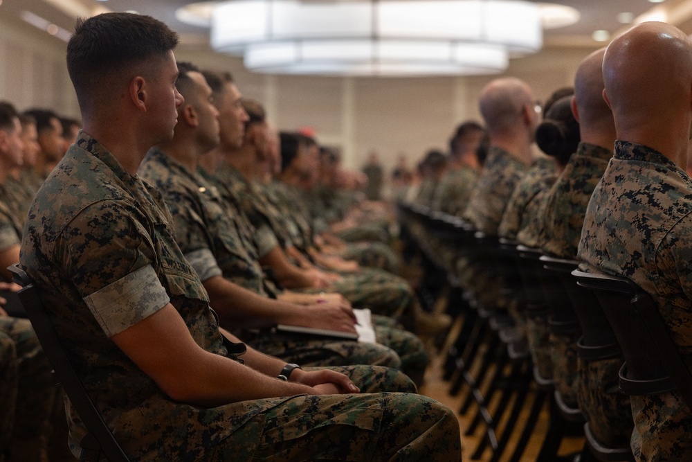 A large group of military personnel in digital camouflage uniforms are seated in rows of chairs inside what appears to be a conference hall or briefing room. The individuals are attentive and facing forward, suggesting they are participating in a formal briefing, training session, or ceremony. The setting is an indoor venue with overhead lighting and a polished floor.