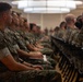 A large group of military personnel in digital camouflage uniforms are seated in rows of chairs inside what appears to be a conference hall or briefing room. The individuals are attentive and facing forward, suggesting they are participating in a formal briefing, training session, or ceremony. The setting is an indoor venue with overhead lighting and a polished floor.