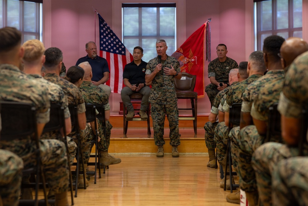 A senior U.S. Marine Corps officer in camouflage utility uniform addresses a formation of Marines seated in rows inside a formal indoor venue. Two civilians and at least one other uniformed Marine are seated on a raised platform behind the speaker. An American flag and a red Marine Corps flag are displayed prominently in the background.