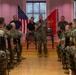A senior U.S. Marine Corps officer in camouflage utility uniform addresses a formation of Marines seated in rows inside a formal indoor venue. Two civilians and at least one other uniformed Marine are seated on a raised platform behind the speaker. An American flag and a red Marine Corps flag are displayed prominently in the background.