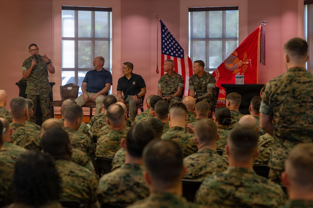 A U.S. Marine Corps briefing or town hall event is taking place in an indoor venue. A panel of individuals, including uniformed Marines and two civilians in casual attire, are seated on a stage while one uniformed Marine speaks into a microphone. A large audience of Marines in camouflage uniforms fills the seating area facing the stage.