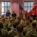A U.S. Marine Corps briefing or town hall event is taking place in an indoor venue. A panel of individuals, including uniformed Marines and two civilians in casual attire, are seated on a stage while one uniformed Marine speaks into a microphone. A large audience of Marines in camouflage uniforms fills the seating area facing the stage.