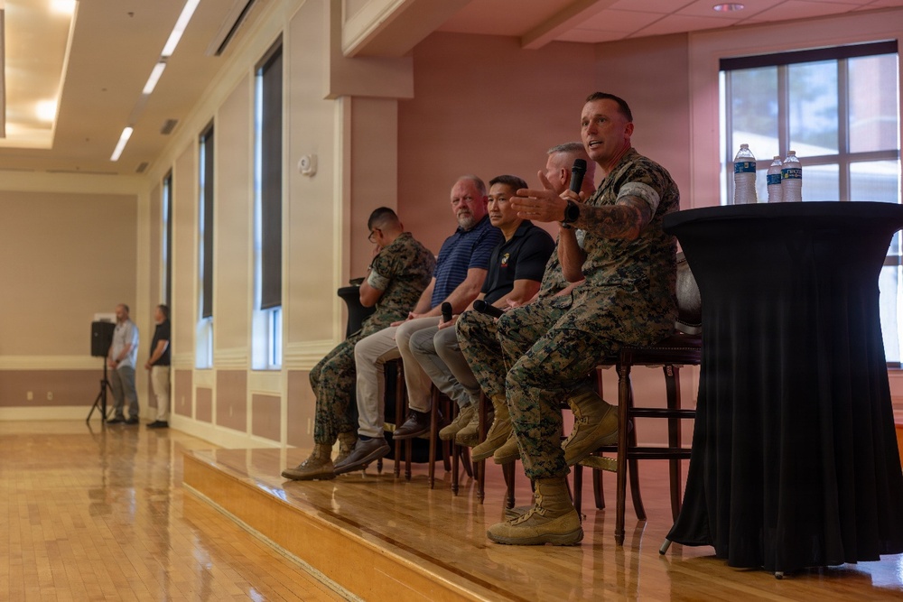 A panel discussion is taking place in what appears to be a military installation event hall or ballroom. Five panelists are seated in a row on a raised platform, including at least three individuals in U.S. Marine Corps camouflage uniforms and two civilians. The rightmost panelist, a Marine, is actively speaking into a microphone with an animated hand gesture.