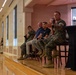 A panel discussion is taking place in what appears to be a military installation event hall or ballroom. Five panelists are seated in a row on a raised platform, including at least three individuals in U.S. Marine Corps camouflage uniforms and two civilians. The rightmost panelist, a Marine, is actively speaking into a microphone with an animated hand gesture.