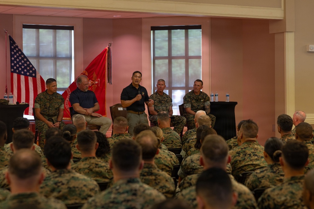 A formal military briefing or town hall is taking place in an indoor venue, with a panel of individuals seated on a stage addressing a large audience of uniformed Marines. One civilian and several military personnel in Marine Corps Combat Utility Uniforms (MCCUU) are seated on the panel, while one individual stands and speaks into a microphone. The audience consists of numerous Marines seated in rows with their backs to the camera.