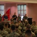 A formal military briefing or town hall is taking place in an indoor venue, with a panel of individuals seated on a stage addressing a large audience of uniformed Marines. One civilian and several military personnel in Marine Corps Combat Utility Uniforms (MCCUU) are seated on the panel, while one individual stands and speaks into a microphone. The audience consists of numerous Marines seated in rows with their backs to the camera.