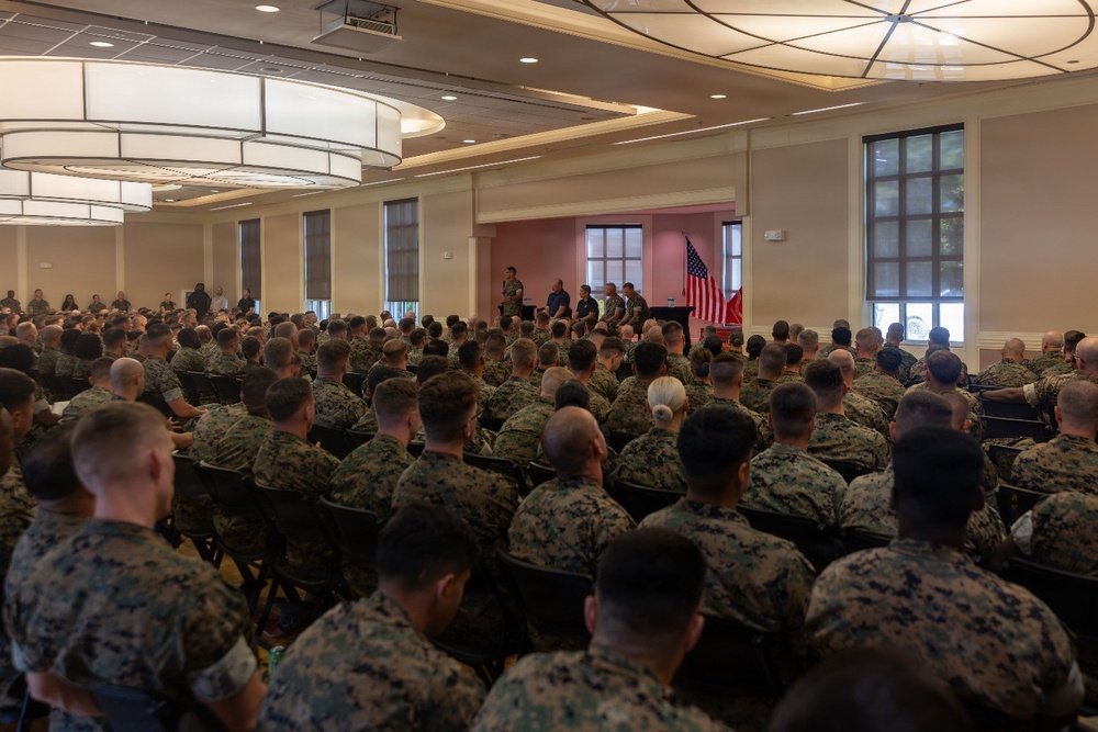 A large indoor military assembly is taking place in a conference or ballroom-style venue, with hundreds of personnel in woodland/digital camouflage uniforms seated in rows of chairs facing a stage. A speaker stands at a podium or microphone at the front of the room, flanked by several other individuals seated on the stage. An American flag and what appears to be a unit or organizational flag are visible at the front of the room.