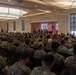 A large indoor military assembly is taking place in a conference or ballroom-style venue, with hundreds of personnel in woodland/digital camouflage uniforms seated in rows of chairs facing a stage. A speaker stands at a podium or microphone at the front of the room, flanked by several other individuals seated on the stage. An American flag and what appears to be a unit or organizational flag are visible at the front of the room.