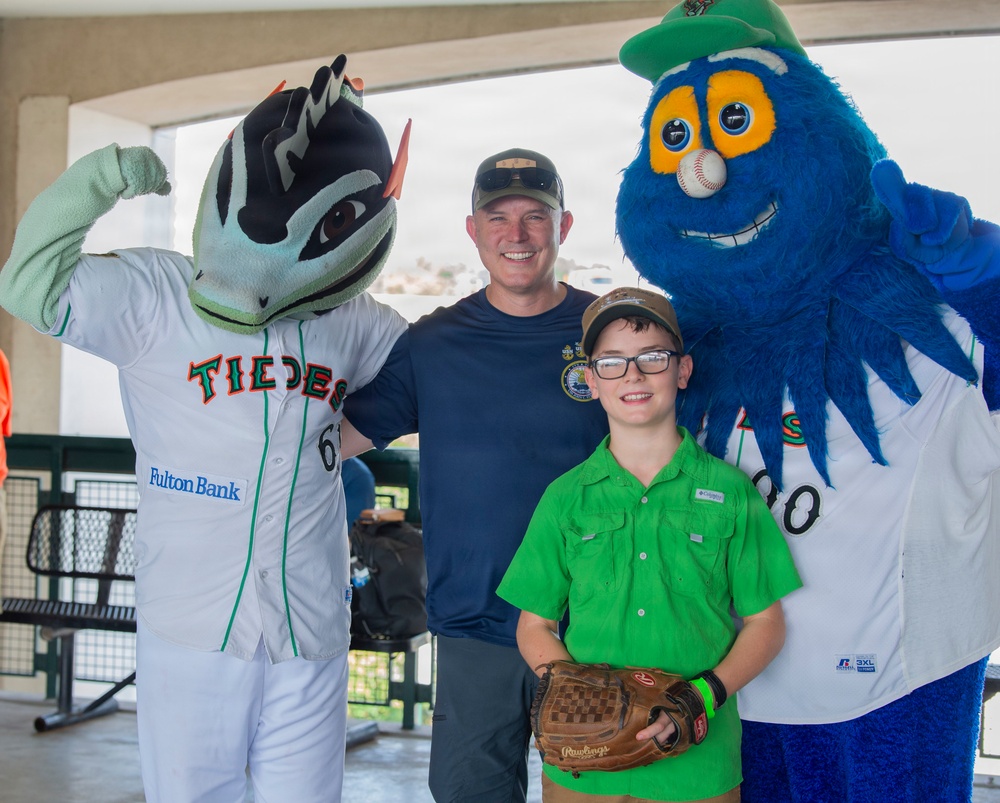 A man and a young boy wearing glasses pose for a photo with two baseball team mascots at what appears to be a Norfolk Tides minor league baseball event. The left mascot wears a 'Tides' jersey with a Fulton Bank sponsor logo, while the right mascot is a large blue fuzzy character with a baseball for a nose. The boy is holding a Rawlings baseball glove, suggesting a fan meet-and-greet or promotional event.