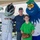 A man and a young boy wearing glasses pose for a photo with two baseball team mascots at what appears to be a Norfolk Tides minor league baseball event. The left mascot wears a 'Tides' jersey with a Fulton Bank sponsor logo, while the right mascot is a large blue fuzzy character with a baseball for a nose. The boy is holding a Rawlings baseball glove, suggesting a fan meet-and-greet or promotional event.