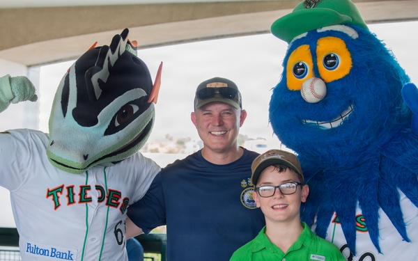 A man and a young boy wearing glasses pose for a photo with two baseball team mascots at what appears to be a Norfolk Tides minor league baseball event. The left mascot wears a 'Tides' jersey with a Fulton Bank sponsor logo, while the right mascot is a large blue fuzzy character with a baseball for a nose. The boy is holding a Rawlings baseball glove, suggesting a fan meet-and-greet or promotional event.