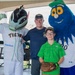 A man and a young boy wearing glasses pose for a photo with two baseball team mascots at what appears to be a Norfolk Tides minor league baseball event. The left mascot wears a 'Tides' jersey with a Fulton Bank sponsor logo, while the right mascot is a large blue fuzzy character with a baseball for a nose. The boy is holding a Rawlings baseball glove, suggesting a fan meet-and-greet or promotional event.