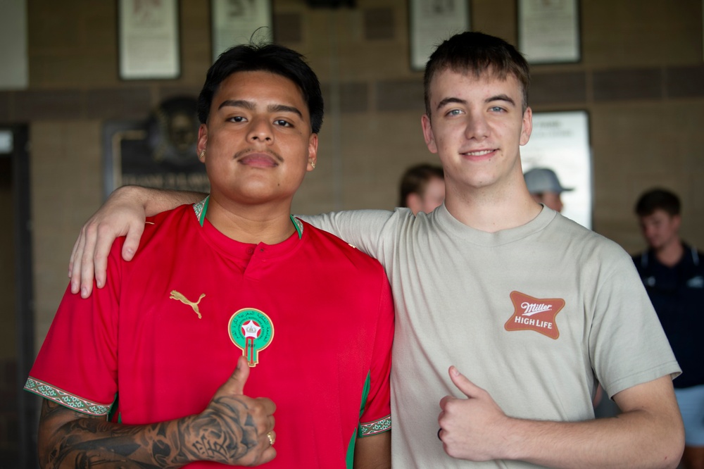 Two young men are posing together for a photo, both giving thumbs up gestures. The person on the left is wearing a red Morocco national football team jersey (Puma) with visible tattoos, while the person on the right is wearing a beige 'Miller High Life' t-shirt. They appear to be in an indoor venue with framed items on the wall in the background.