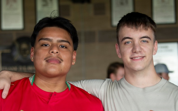Two young men are posing together for a photo, both giving thumbs up gestures. The person on the left is wearing a red Morocco national football team jersey (Puma) with visible tattoos, while the person on the right is wearing a beige 'Miller High Life' t-shirt. They appear to be in an indoor venue with framed items on the wall in the background.