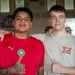 Two young men are posing together for a photo, both giving thumbs up gestures. The person on the left is wearing a red Morocco national football team jersey (Puma) with visible tattoos, while the person on the right is wearing a beige 'Miller High Life' t-shirt. They appear to be in an indoor venue with framed items on the wall in the background.