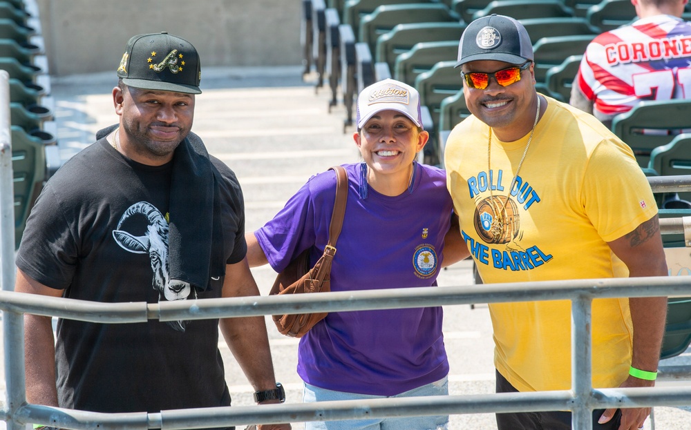 Three people pose together for a photo in what appears to be a stadium or sports venue with green seats visible in the background. They are smiling and appear to be fans attending a sporting event on a sunny day. A fourth person wearing a jersey with 'CORONE' visible is partially seen in the background.