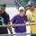 Three people pose together for a photo in what appears to be a stadium or sports venue with green seats visible in the background. They are smiling and appear to be fans attending a sporting event on a sunny day. A fourth person wearing a jersey with 'CORONE' visible is partially seen in the background.