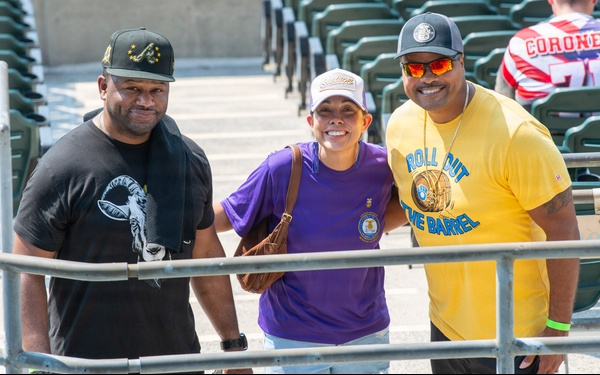 Three people pose together for a photo in what appears to be a stadium or sports venue with green seats visible in the background. They are smiling and appear to be fans attending a sporting event on a sunny day. A fourth person wearing a jersey with 'CORONE' visible is partially seen in the background.