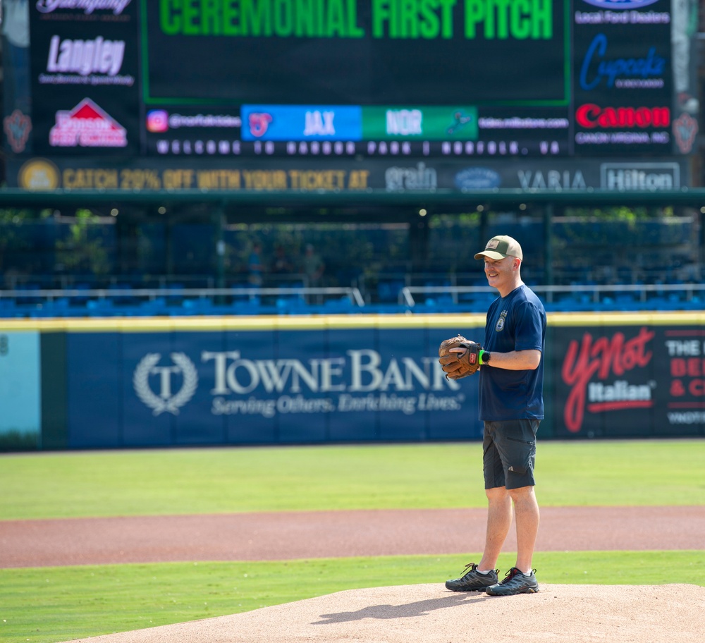 A man stands on a baseball pitcher's mound holding a glove, preparing for or having just completed a ceremonial first pitch at Harbor Park in Norfolk, Virginia. The scoreboard behind him displays 'CEREMONIAL FIRST PITCH' with a matchup between JAX and NOR. The stadium features multiple sponsor advertisements including TowneBank, Canon, Hilton, and YNot Italian.