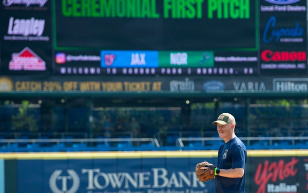 A man stands on a baseball pitcher's mound holding a glove, preparing for or having just completed a ceremonial first pitch at Harbor Park in Norfolk, Virginia. The scoreboard behind him displays 'CEREMONIAL FIRST PITCH' with a matchup between JAX and NOR. The stadium features multiple sponsor advertisements including TowneBank, Canon, Hilton, and YNot Italian.