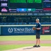 A man stands on a baseball pitcher's mound holding a glove, preparing for or having just completed a ceremonial first pitch at Harbor Park in Norfolk, Virginia. The scoreboard behind him displays 'CEREMONIAL FIRST PITCH' with a matchup between JAX and NOR. The stadium features multiple sponsor advertisements including TowneBank, Canon, Hilton, and YNot Italian.