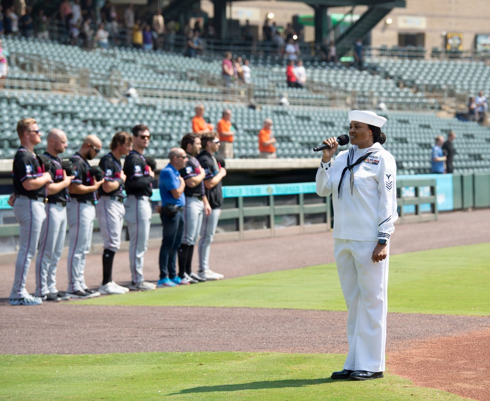 A U.S. Navy sailor in dress white uniform is singing into a microphone, likely performing the National Anthem, at a minor league baseball stadium. A line of baseball players in dark jerseys and gray pants stand at attention along the baseline. The event appears to be a pre-game ceremony on a sunny day with a partially filled stadium in the background.