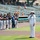 A U.S. Navy sailor in dress white uniform is singing into a microphone, likely performing the National Anthem, at a minor league baseball stadium. A line of baseball players in dark jerseys and gray pants stand at attention along the baseline. The event appears to be a pre-game ceremony on a sunny day with a partially filled stadium in the background.