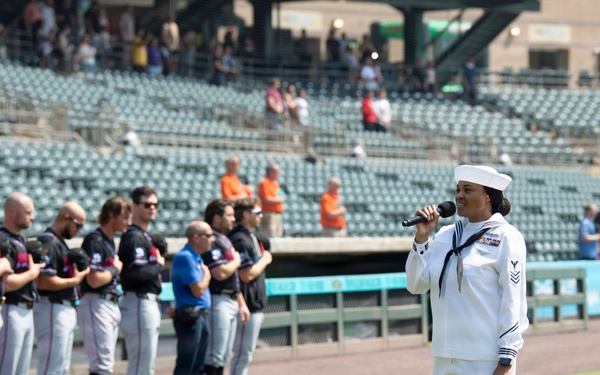 A U.S. Navy sailor in dress white uniform is singing into a microphone, likely performing the National Anthem, at a minor league baseball stadium. A line of baseball players in dark jerseys and gray pants stand at attention along the baseline. The event appears to be a pre-game ceremony on a sunny day with a partially filled stadium in the background.