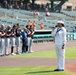 A U.S. Navy sailor in dress white uniform is singing into a microphone, likely performing the National Anthem, at a minor league baseball stadium. A line of baseball players in dark jerseys and gray pants stand at attention along the baseline. The event appears to be a pre-game ceremony on a sunny day with a partially filled stadium in the background.