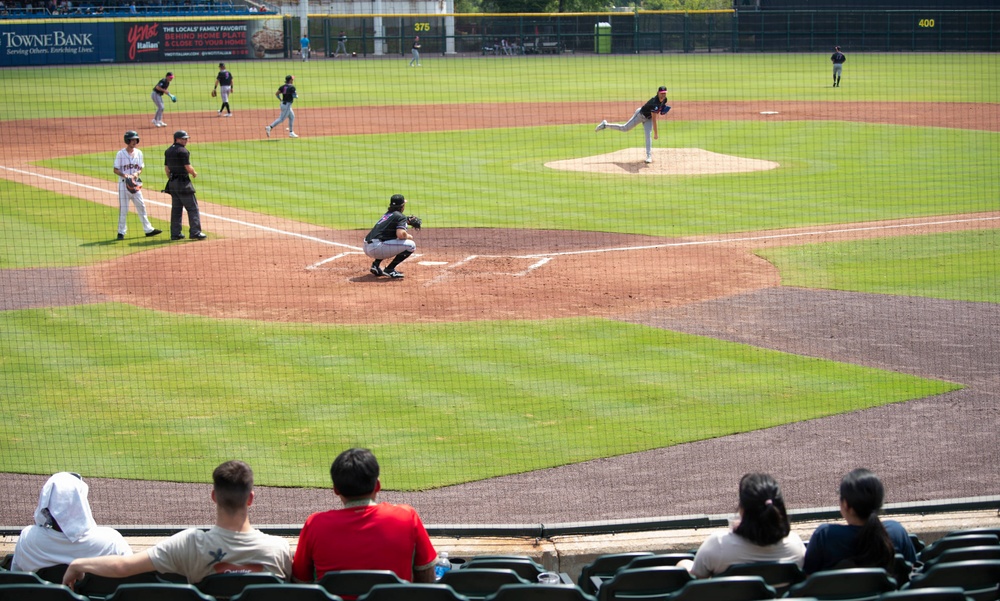 A baseball game is in progress at a minor league or collegiate stadium, captured from behind home plate in the stands. A pitcher is mid-delivery on the mound while a catcher crouches in position at home plate, with fielders visible throughout the infield and outfield. A small group of spectators watches from the nearly empty stands in the foreground.