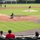 A baseball game is in progress at a minor league or collegiate stadium, captured from behind home plate in the stands. A pitcher is mid-delivery on the mound while a catcher crouches in position at home plate, with fielders visible throughout the infield and outfield. A small group of spectators watches from the nearly empty stands in the foreground.