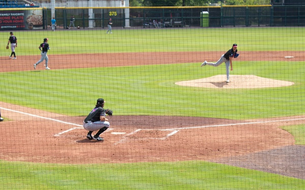 A baseball game is in progress at a minor league or collegiate stadium, captured from behind home plate in the stands. A pitcher is mid-delivery on the mound while a catcher crouches in position at home plate, with fielders visible throughout the infield and outfield. A small group of spectators watches from the nearly empty stands in the foreground.