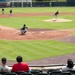 A baseball game is in progress at a minor league or collegiate stadium, captured from behind home plate in the stands. A pitcher is mid-delivery on the mound while a catcher crouches in position at home plate, with fielders visible throughout the infield and outfield. A small group of spectators watches from the nearly empty stands in the foreground.