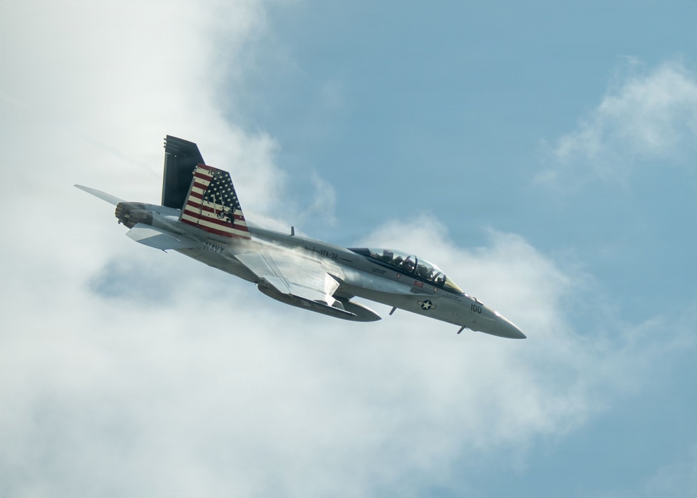 A U.S. Navy F/A-18 Super Hornet jet fighter is captured in flight against a partly cloudy blue sky. The aircraft features a distinctive American flag livery on its tail fins and bears the number '100' on its nose. The aircraft appears to be performing a high-speed maneuver, with visible vapor condensation forming along the fuselage.