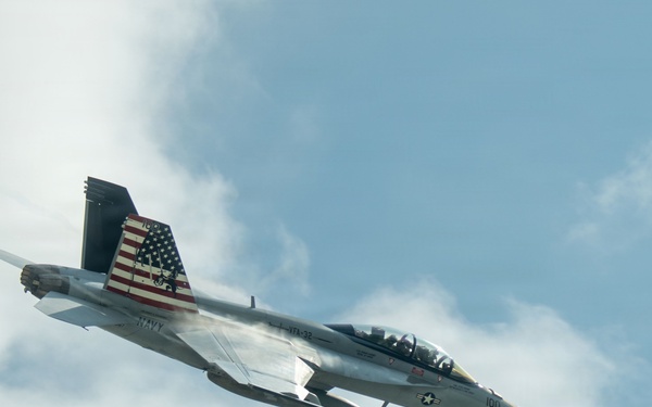 A U.S. Navy F/A-18 Super Hornet jet fighter is captured in flight against a partly cloudy blue sky. The aircraft features a distinctive American flag livery on its tail fins and bears the number '100' on its nose. The aircraft appears to be performing a high-speed maneuver, with visible vapor condensation forming along the fuselage.