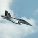 A U.S. Navy F/A-18 Super Hornet jet fighter is captured in flight against a partly cloudy blue sky. The aircraft features a distinctive American flag livery on its tail fins and bears the number '100' on its nose. The aircraft appears to be performing a high-speed maneuver, with visible vapor condensation forming along the fuselage.